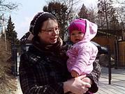 Mutter hält Baby in handgemachter Winterjacke und Mütze, fröhliches Familienbild bei Spaziergang im Freien, Fokus auf handgemachte Kleidung und Naturumgebung.