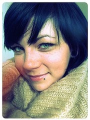 Nahaufnahme einer jungen Frau mit dunklem Kurzhaarschnitt, blauen Augen, Sommersprossen und Lippenpiercing, die einen warmen Schal trägt.