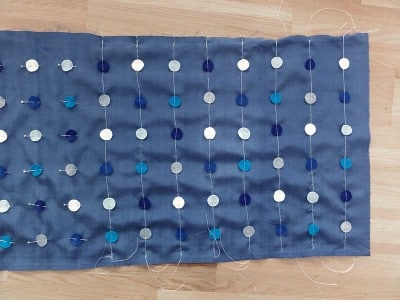 Handgefertigter blauer Seidenschal mit vertikalen Reihen von weißen, hellblauen und dunklen blauen Perlen, die an dünnen Fäden befestigt sind, auf einem Holzuntergrund.