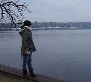 Frau steht am Ufer eines Flusses, blickt nachdenklich auf das Wasser, trägt eine Jacke, Jeans und Stiefel, im Hintergrund sind Bäume und eine Stadt sichtbar.