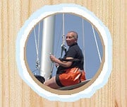 Mann sitzt auf einem Segelboot, hält das Steuerrad, mit einem blauen Himmel im Hintergrund, ideal für maritime und Segelthemen.