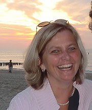 Lachende Frau am Strand bei Sonnenuntergang, mit Meer, Himmel und Sonnenstrahlen im Hintergrund, trägt Perlenkette und Sonnenbrille auf Kopf.
