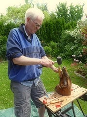 Älterer Mann beim Holzschnitzen im Garten, umgeben von blühenden Pflanzen und Bäumen, zeigt handwerkliches Können bei einem kreativen DIY-Projekt.