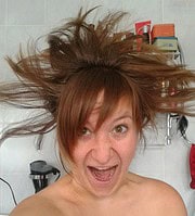 Frau mit wild aufgestellten, zerzausten Haaren, lachend und fröhlich, in einem kreativen Umfeld mit Handwerksmaterialien im Hintergrund, symbolisiert kreative Handarbeit und Spaß.