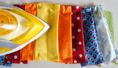 Verschiedene bunte Stoffe in Reihen, bereit für das Nähen, mit einem Bügeleisen im Vordergrund, ideal für kreative Näharbeiten bei HANDMADE Kultur.
