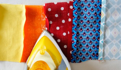 Verschiedene bunte Stoffe in Gelb, Orange, Rot, Blau und Hellblau für Näharbeiten, mit Bügeleisen im Vordergrund, geeignet für Sew Along Tag 3: Plissee und Innentasche nähen.