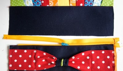 Bunte Stoffe und Reißverschlüsse für eine selbstgenähte Clutch, inklusive einer roten Schleife mit weißen Punkten, ideal für den Sew Along Tag 4.