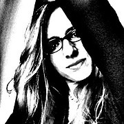 Frau mit Brille und langen Haaren, lächelnd, in einem kreativen Handwerksumfeld, symbolisiert handgemachte Kunst und kreative Kultur.