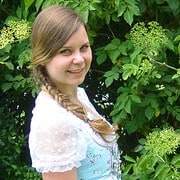 Junge Frau mit Zopf und weißem Kleid vor üppigem grünen Laub und Blüten, lächelnd, natürliche Umgebung, sommerlich, entspannt, authentisch, fröhlich, Naturfotografie.