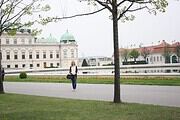 Frau mit Rucksack spaziert im Park vor historischen Gebäuden, umgeben von Bäumen und grüner Wiese, bei ruhigem Wetter in einer Stadt.