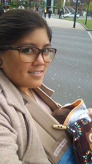 Frau mit Brille und Strickjacke hält ein handgemachtes, bunt besticktes Stofftier, im Hintergrund eine Stadtstraße mit Fußgängern und Autos.