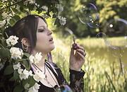 Frau mit dunklem Haar und Piercings bläst Seifenblasen in einer Naturkulisse mit Blüten und Bäumen, kreative Handwerkskunst und Naturverbundenheit im Fokus.