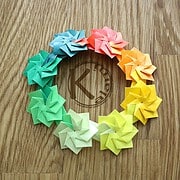 Bunter Origami-Blumenkranz aus Papier in Regenbogenfarben, handgefertigt, auf Holztisch gelegt, kreative Bastelarbeit für Dekoration oder Geschenk.