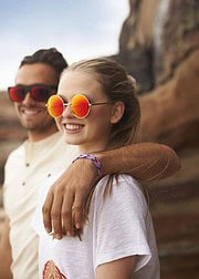 Junge Frau trägt Makraméband mit bunten Perlen, lächelnd, mit Freund im Hintergrund, bei sonnigem Wetter, ideal für Sommermode, handgefertigt, modisch, Accessoire, Trend.