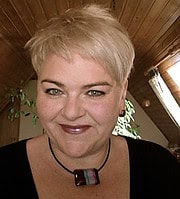 Porträt einer Frau mit kurzen blonden Haaren, dunklem Oberteil und Schmuck, in einem Holzhaus mit Pflanzen im Hintergrund, bei Handmade Kultur.