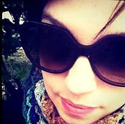 Frau trägt große schwarze Sonnenbrille, trägt bunte Kleidung und hat braune Haare, die leicht im Wind wehen, umgeben von Natur, ideal für Mode- und Lifestyle-Bilder bei HANDMADE Kultur.
