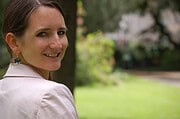 Frau mit braunen Haaren und weißen Jacke lächelt in einem Park mit grünen Bäumen und Rasen, symbolisiert kreative Handwerkskunst und Naturverbundenheit.