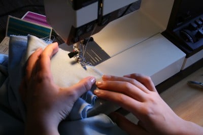 Hand beim Nähen eines Polsters mit Nähmaschine, Stoffe und Nähutensilien sichtbar, im Zusammenhang mit Polsterarbeiten und Handarbeit.