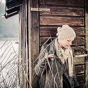 Frau in Winterkleidung mit Strickmütze und Schal vor einer Holzfassade, umgeben von Natur, zeigt handgemachte Kleidung und Accessoires, die für Handmade Kultur stehen.