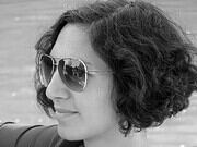 Frau mit lockigem Haar und Sonnenbrille, die entspannt am Wasser sitzt, bei Handmade Kultur, Fokus auf handgemachte Mode und Accessoires.