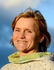 Porträt einer lächelnden Frau mit kurzen, hellbraunen Haaren, die eine grüne Halstuch trägt, vor einem blauen Himmel, symbolisiert kreative Handarbeit und Kunsthandwerk.