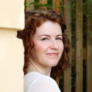 Porträt einer lächelnden Frau mit roten Locken, die an einer gelben Wand steht, umgeben von grünen Pflanzen, im Kontext von HANDMADE Kultur.