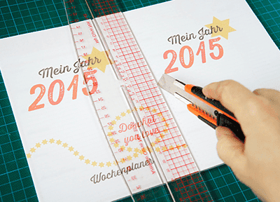 Hand schneidet einen Wochenkalender für 2015 mit einem Cuttermesser aus, auf einem Schneidebrett mit Lineal, für kreative Handarbeit und Kalenderplanung.