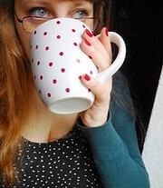 Frau mit Brille trinkt aus einem weißen Keramikbecher mit roten Punkten, die Hand mit roten Nagellack hält den Becher, im Hintergrund unscharf, Fokus auf den Becher.