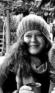 Frau mit lockigem Haar, gestrickter Mütze und Schal, hält eine Tasse Kaffee, umgeben von handgemachten Dekorationen und Pflanzen, bei Handmade Kultur Veranstaltung.