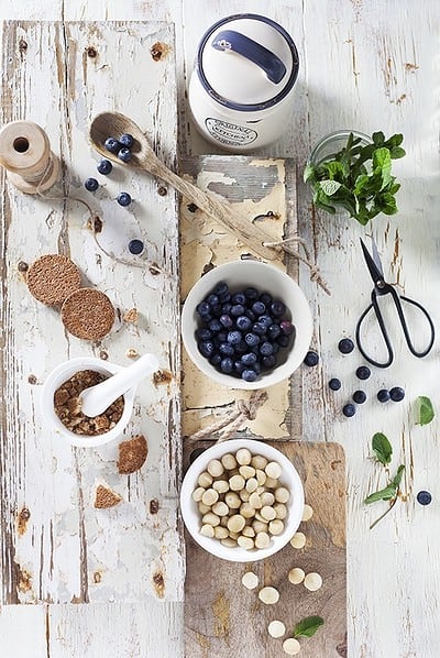 Frische Blaubeeren, weiße Schokoladenkugeln, Minze und Küchenutensilien auf weißem Holztablett, ideal für Back- und Dessertrezepte.