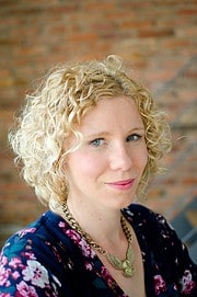 Porträt einer Frau mit lockigem blondem Haar, blauer Bluse mit Blumenmuster, Schmuck, vor einer Backsteinwand, Fokus auf Gesicht, natürliche Beleuchtung, kreative Handwerkskunst.