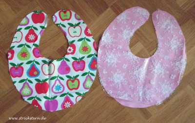 Zwei handgemachte Baby-Lätzchen, eines mit Apfelmuster in Rot, Grün und Pink, das andere in zartem Rosa mit weißen Blumen, ideal als Last-Minute Babygeschenk.