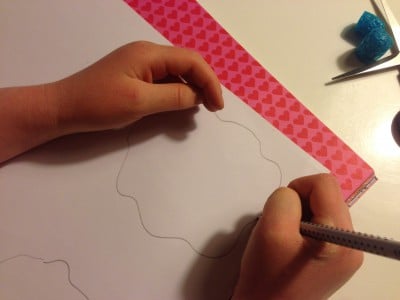 Hand beim Zeichnen einer Kontur für eine Papierblume, Bastelmaterialien wie blauer Schwamm und Schere sichtbar, kreative Frühlingsdekoration.