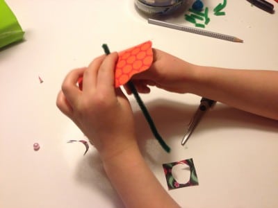 Hand beim Basteln einer Papierblume mit orangen Blütenblatt und grüner Stiel, kreative Handarbeit für Frühling und Handmadekultur.