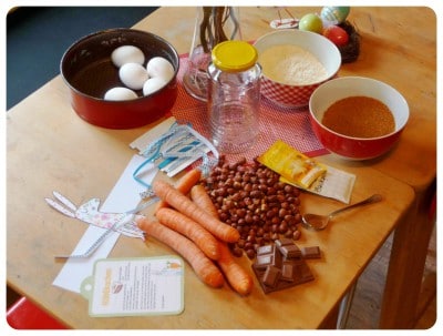 Osterdekoration mit bunten Eiern, Schokolade, Karotten, Nüssen, Mehl, Zucker, und einem Glas Wasser auf einem Holztisch, passend für Frühlings- und Osterfeierlichkeiten.