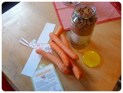Frische Karotten, ein Glas mit Möhrenkuchen, ein Löffel Honig und eine Karte auf einem Holztisch, passend zum Thema "Hattu Möhrchen?" - Möhrenkuchen-Backmischung im Glas.
