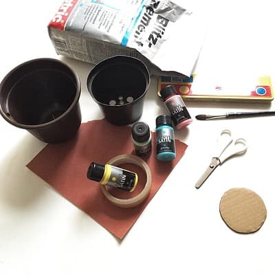 Selbstgemachter Zement-Übertopf mit Bastelmaterialien, Farben, Pinsel, Schere und Tonpapier für kreative Blumenarrangements.