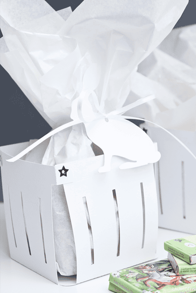 Weißes Osterkörbchen aus Pappe mit Geschenkpapier und Hasenanhänger, ideal für Ostergeschenke und Dekorationen.