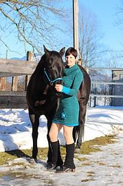 Frau mit Kurzhaarfrisur in türkisfarbenem Kleid und schwarzen Stiefeln steht neben einem schwarzen Pferd im Schnee, umgeben von Winterlandschaft und Holzzaun.