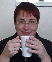 Frau mit Brille und Kurzhaarfrisur hält eine weiße Tasse und lächelt in die Kamera, im Hintergrund eine weiße Wand mit Rahmen.
