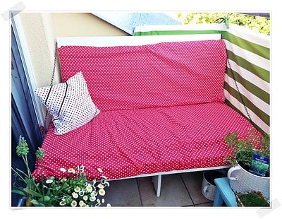Balkon-Couch mit rot-weiß gepunkteten Kissen und grün-weiß gestreifter Seitenbespannung, umgeben von Pflanzen, ideal für entspannte Stunden im Freien.