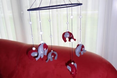 Handgefertigtes Elefanten-Mobile aus Stoff in Rot und Grau, das an einer schwarzen Stange über einem roten Sofa hängt, ideal für Babyzimmer.