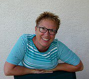 Lächelnde Frau mit Brille und kurzen Haaren, trägt ein blau-weiß gestreiftes Shirt, sitzt vor weißer Wand, vermittelt Kreativität und Handwerkskunst.