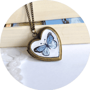 Herzförmiger Anhänger mit einem blauen Schmetterling im Inneren, an einer Kette, auf Holz und Stoff liegend, für handgemachte Schmuckkunst und kreative Accessoires.