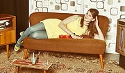 Frau in gelbem Kleid liegt entspannt auf einem braunen Sofa, umgeben von Vintage-Möbeln und einer gemütlichen Wohnatmosphäre, ideal für Handmade- und Kulturthemen.