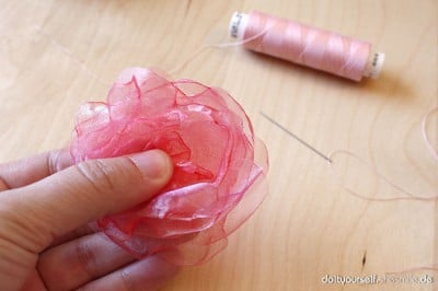 Hand hält eine Blume aus rosa Organza-Stoff, daneben eine Spule mit rosa Nähgarn auf einem Holzarbeitsplatz.