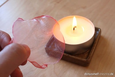 Herzförmiges Organza-Stück wird neben einer brennenden Kerze auf Holzuntergrund gehalten, geeignet für Bastel- und Dekorationsideen.