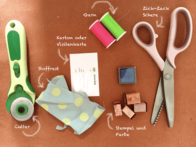 Bastelmaterialien für Karten: Cutter, Stempel, Farben, Garn, Schere, Papier, Stempel und Farbe, Zick-Zack Schere, Karton oder Visitenkarte, Stoffrest, Zuck-Zack Garn, Stempel.