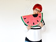 Frau mit roten Haaren und Brille hält ein großes, buntes Wassermelonen-Kissen, das als Dekoration oder Kuscheltier dient, vor weißer Wand mit Schriftzug.