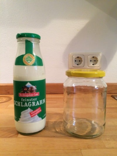 Glas mit Sahne und leeres Einmachglas auf Holzoberfläche, Steckdose im Hintergrund, für DIY Butterherstellung.